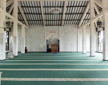 A spacious interior of a mosque with rows of green carpet, arched white marble walls, and tall pillars. The ceiling is vaulted with wooden beams, and there is a wooden podium at the front. There are people seen praying, and a digital clock on the wall shows the time. A hanging chandelier and a speaker are visible.