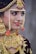 A person is adorned in elaborate traditional jewelry, featuring gold, red, and pearl elements. The jewelry includes a detailed headpiece, large earrings, a nose ring, and a heavily decorated necklace.