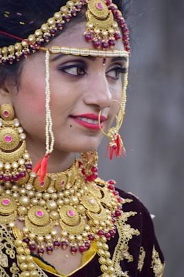 A person is adorned in elaborate traditional jewelry, featuring gold, red, and pearl elements. The jewelry includes a detailed headpiece, large earrings, a nose ring, and a heavily decorated necklace.