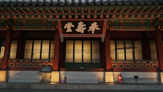A traditional Korean building with ornate wooden architecture, featuring a large sign with intricate Korean calligraphy. The wooden structure includes colorful decorative elements and lattice windows. A fire extinguisher is visible on the stone steps in front.
