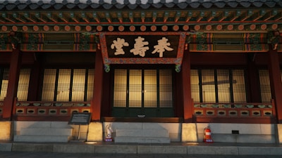A traditional Korean building with ornate wooden architecture, featuring a large sign with intricate Korean calligraphy. The wooden structure includes colorful decorative elements and lattice windows. A fire extinguisher is visible on the stone steps in front.