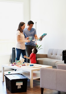 A family scene with two adults and a child in a living room. The adults are standing near a table holding a board game, and the child is reaching up to interact with them. The room is furnished with a sofa, a table with toys, and a large black storage box.