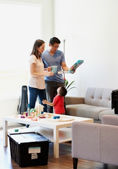 A family scene with two adults and a child in a living room. The adults are standing near a table holding a board game, and the child is reaching up to interact with them. The room is furnished with a sofa, a table with toys, and a large black storage box.