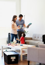 A family scene with two adults and a child in a living room. The adults are standing near a table holding a board game, and the child is reaching up to interact with them. The room is furnished with a sofa, a table with toys, and a large black storage box.