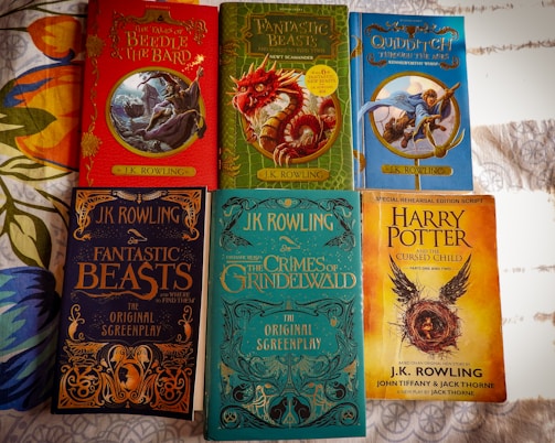 Several books written by J.K. Rowling are arranged on a surface. The covers display colorful illustrations and titles such as 'The Tales of Beedle the Bard', 'Fantastic Beasts and Where to Find Them', 'Quidditch Through the Ages', 'Fantastic Beasts: The Crimes of Grindelwald', and 'Harry Potter and the Cursed Child'. Each book has a distinct cover design featuring magical themes and characters.
