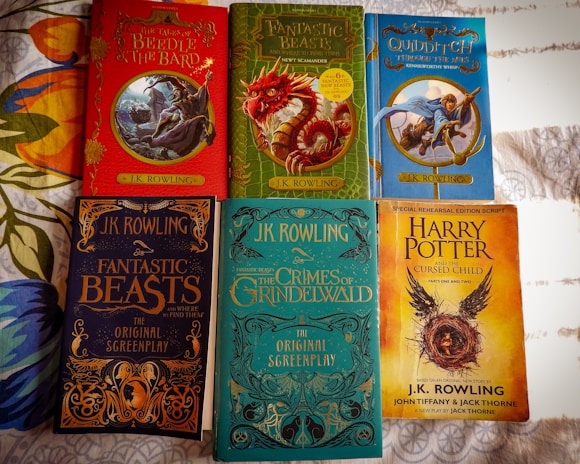 Several books written by J.K. Rowling are arranged on a surface. The covers display colorful illustrations and titles such as 'The Tales of Beedle the Bard', 'Fantastic Beasts and Where to Find Them', 'Quidditch Through the Ages', 'Fantastic Beasts: The Crimes of Grindelwald', and 'Harry Potter and the Cursed Child'. Each book has a distinct cover design featuring magical themes and characters.