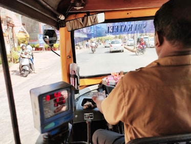 The view from inside a rickshaw, showing the driver in a brown uniform steering the vehicle down a busy urban street. The meter in the foreground displays some digits, and various vehicles, including scooters and cars, are visible through the windshield. A mirror reflects part of the street scene, and decorations are placed on the dashboard.