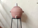 A large clay water storage pot with a metal lid is positioned on a black metal stand. The pot is equipped with a small tap at the bottom.