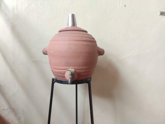 A large clay water storage pot with a metal lid is positioned on a black metal stand. The pot is equipped with a small tap at the bottom.