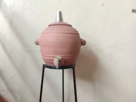 A large clay water storage pot with a metal lid is positioned on a black metal stand. The pot is equipped with a small tap at the bottom.