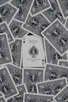 A collection of playing cards spread out, with an ace of spades card prominently displayed in the center. The cards have intricate black and white designs featuring ornate patterns and figures.
