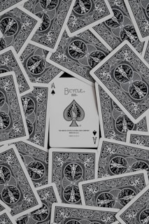 A collection of playing cards spread out, with an ace of spades card prominently displayed in the center. The cards have intricate black and white designs featuring ornate patterns and figures.