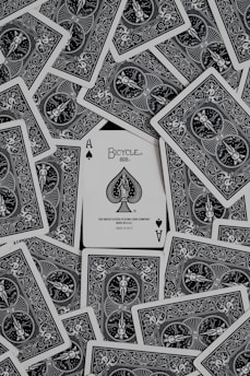 A collection of playing cards spread out, with an ace of spades card prominently displayed in the center. The cards have intricate black and white designs featuring ornate patterns and figures.