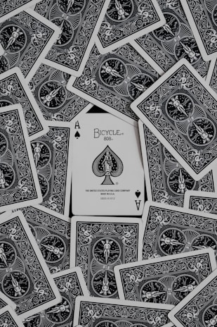 A collection of playing cards spread out, with an ace of spades card prominently displayed in the center. The cards have intricate black and white designs featuring ornate patterns and figures.