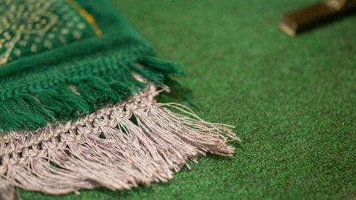 A close-up view of a woven textile with fringed edges. The fabric features a rich green color with textured detailing, including intricate patterns and stitching. The edges are adorned with grey tassels that add a tactile element to the design. The background appears to be a solid green surface, providing contrast to the fabric.