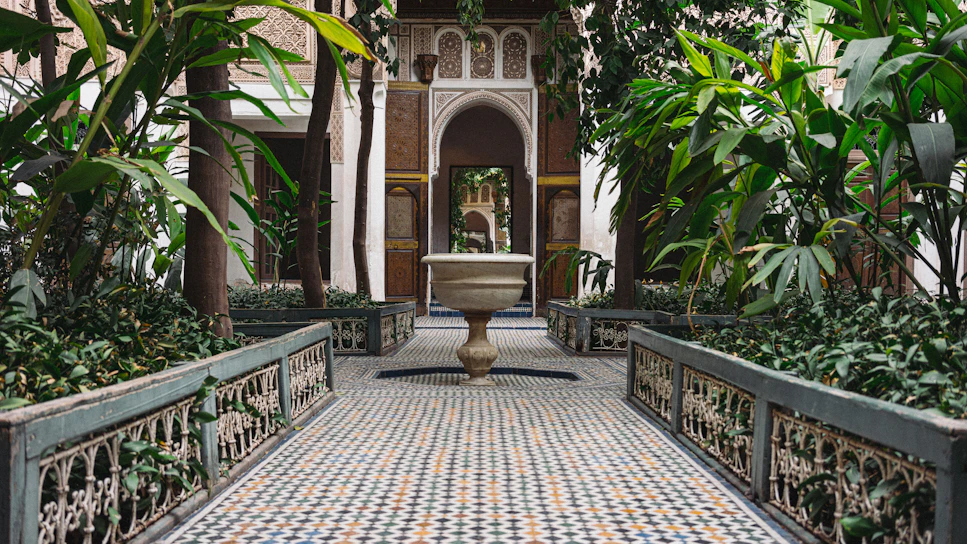 A sunlit Moroccan courtyard with lush greenery and traditional geometric tile patterns on the walls.