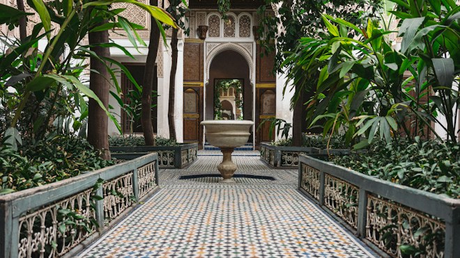 An intricately tiled courtyard with symmetrical mosaic patterns on the floor, surrounded by lush green plants and ornate architectural elements. The focal point is a decorative stone fountain centered in the scene, with arches and detailed wall designs in the background.