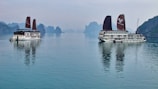 Two traditional-style cruise ships with brown sails are positioned on a calm body of water, with reflections visible on the surface. In the background, karst limestone formations and a hazy sky create a serene landscape.
