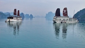 Two traditional-style cruise ships with brown sails are positioned on a calm body of water, with reflections visible on the surface. In the background, karst limestone formations and a hazy sky create a serene landscape.