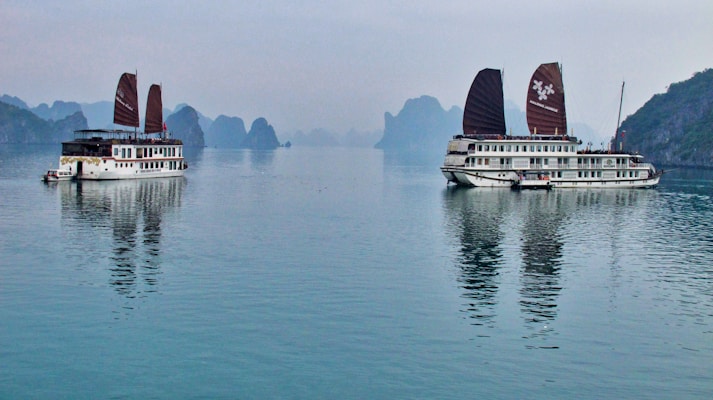 Two traditional-style cruise ships with brown sails are positioned on a calm body of water, with reflections visible on the surface. In the background, karst limestone formations and a hazy sky create a serene landscape.