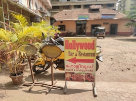 A signboard for 'Hollywood Bar & Restaurant' is situated on a sidewalk next to potted plants and some large spoons. The sign is yellow with large red text and contains images of food. In the background, there is a small building with a red tiled roof, a few parked motorcycles, and an auto rickshaw.