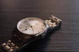 A close-up view of a luxury wristwatch with a metallic bracelet laying on a dark textured surface. The watch has a round face with a silver bezel, a white dial, and minimalist hour markers. The lighting creates subtle reflections on the metal, enhancing the elegant appearance of the timepiece.