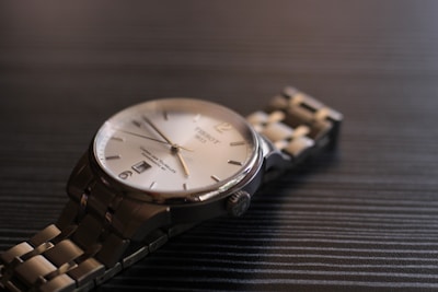 A close-up view of a luxury wristwatch with a metallic bracelet laying on a dark textured surface. The watch has a round face with a silver bezel, a white dial, and minimalist hour markers. The lighting creates subtle reflections on the metal, enhancing the elegant appearance of the timepiece.