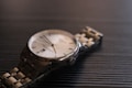 A close-up view of a luxury wristwatch with a metallic bracelet laying on a dark textured surface. The watch has a round face with a silver bezel, a white dial, and minimalist hour markers. The lighting creates subtle reflections on the metal, enhancing the elegant appearance of the timepiece.