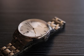 A close-up view of a luxury wristwatch with a metallic bracelet laying on a dark textured surface. The watch has a round face with a silver bezel, a white dial, and minimalist hour markers. The lighting creates subtle reflections on the metal, enhancing the elegant appearance of the timepiece.