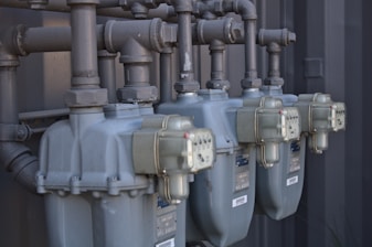 Several industrial gas meters and pipes are mounted on a metal structure. The meters are arranged in a row and connected to a network of thick pipes above. The overall industrial setting appears organized and functional.
