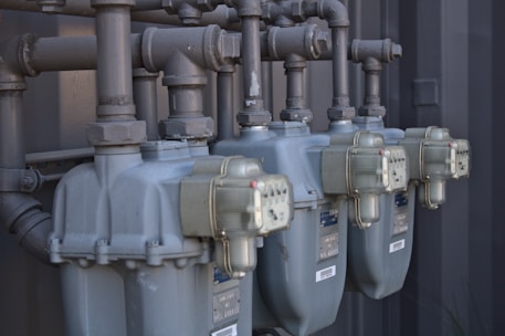 Several industrial gas meters and pipes are mounted on a metal structure. The meters are arranged in a row and connected to a network of thick pipes above. The overall industrial setting appears organized and functional.