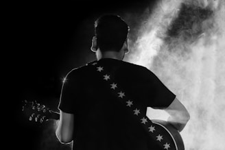 A person is playing a guitar with their back facing the viewer. The scene is dramatic with heavy smoke or mist surrounding the performer. The guitar strap has a pattern of stars on it. The image is in black and white, enhancing the moody and atmospheric feel.