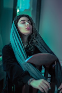 A woman with closed eyes and serene expression is seated on a wooden chair. She wears a dark coat and has a scarf draped over her head. The lighting casts a blue-green hue over the scene, contributing to a calming ambiance.