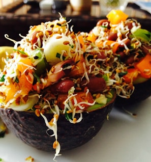 A vibrant salad is served inside avocado shells, featuring a mix of sprouts, beans, diced tomatoes, cucumbers, and topped with seeds.