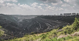 A large quarry or open-pit mine stretches across the landscape, with terraced levels and winding paths. The scene is bordered by green vegetation in the foreground, while an industrial cityscape with buildings and smokestacks is visible on the horizon under a cloud-filled sky.