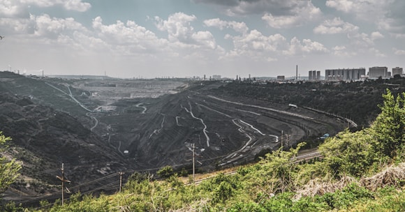 A large quarry or open-pit mine stretches across the landscape, with terraced levels and winding paths. The scene is bordered by green vegetation in the foreground, while an industrial cityscape with buildings and smokestacks is visible on the horizon under a cloud-filled sky.