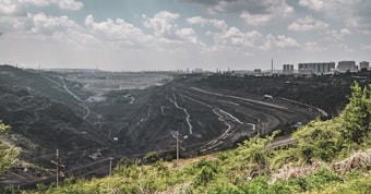 A large quarry or open-pit mine stretches across the landscape, with terraced levels and winding paths. The scene is bordered by green vegetation in the foreground, while an industrial cityscape with buildings and smokestacks is visible on the horizon under a cloud-filled sky.