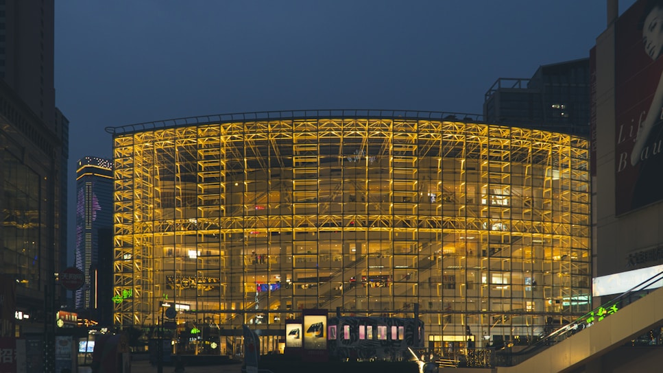 A large, modern building with a distinctive glass facade illuminated in warm yellow lights. The structure appears to be a shopping center or commercial space, with visible advertisements and shopfronts. Surrounding it are tall skyscrapers, adding to the urban landscape. The scene takes place in the evening with a backdrop of a darkening sky.