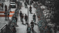 A row of bicycles is lined up along a sidewalk, with several people walking and riding bikes or scooters. The scene has an urban setting and appears to be in a city where bike-sharing is common. People are casually dressed, and some are wearing masks, reflecting a contemporary, everyday atmosphere.