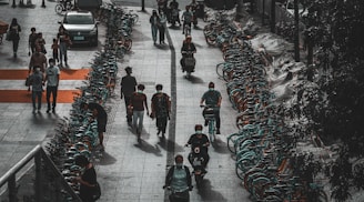 A row of bicycles is lined up along a sidewalk, with several people walking and riding bikes or scooters. The scene has an urban setting and appears to be in a city where bike-sharing is common. People are casually dressed, and some are wearing masks, reflecting a contemporary, everyday atmosphere.