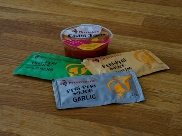A selection of Nando's sauce packets and a small tub of Nando's Chilli Jam are placed on a wooden surface. The sauce packets include flavors such as Wild Herb, Medium, and Garlic in colorful packaging.
