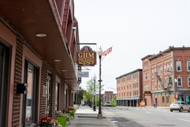 A quiet street scene features a row of brick buildings, including the GHM Insurance Agency. The street is lined with sidewalks adorned with colorful flowers in planters. An American flag is mounted on a lamppost, and across the street are more brick buildings with architectural details. A few cars are parked along the street.