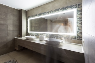 A modern bathroom features gray tiled walls and a long wooden counter with two white vessel sinks. An illuminated rectangular mirror with geometric patterned borders is positioned above the sinks. A blurred figure is partially visible, creating a ghost-like effect.