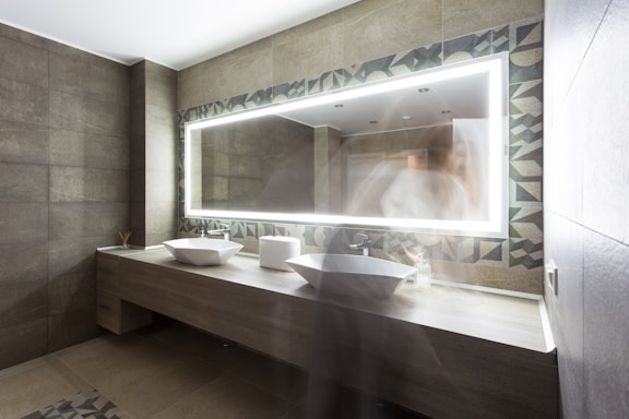 A modern bathroom features gray tiled walls and a long wooden counter with two white vessel sinks. An illuminated rectangular mirror with geometric patterned borders is positioned above the sinks. A blurred figure is partially visible, creating a ghost-like effect.