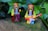 Two LEGO figurines dressed in hippie-style clothing are placed outdoors against a backdrop of green leaves. One figure is holding a black camera, while the other holds a red electric guitar.