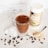 A glass mug filled with a chocolate beverage topped with cocoa powder, placed on a marble surface. Ice cubes and coffee beans are scattered around the mug. Nearby, a spoon with protein powder and a container of protein powder are visible.