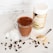 A glass mug filled with a chocolate beverage topped with cocoa powder, placed on a marble surface. Ice cubes and coffee beans are scattered around the mug. Nearby, a spoon with protein powder and a container of protein powder are visible.
