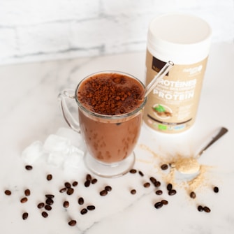 A glass mug filled with a chocolate beverage topped with cocoa powder, placed on a marble surface. Ice cubes and coffee beans are scattered around the mug. Nearby, a spoon with protein powder and a container of protein powder are visible.