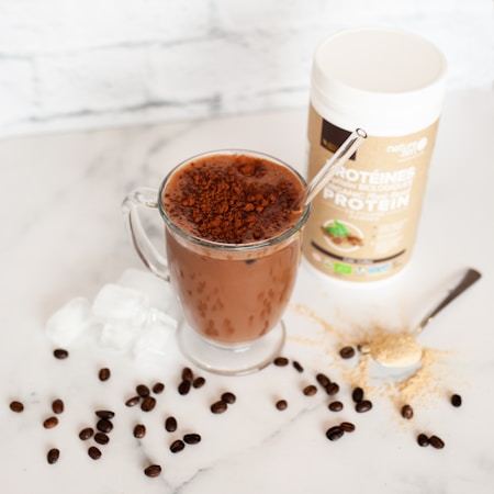 A glass mug filled with a chocolate beverage topped with cocoa powder, placed on a marble surface. Ice cubes and coffee beans are scattered around the mug. Nearby, a spoon with protein powder and a container of protein powder are visible.