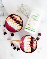 Two smoothie bowls filled with a pinkish berry blend are topped with sliced bananas, cherries, almond slices, and shredded coconut. A wooden spoon lies beside the bowls. Scattered cherries and a container of organic protein powder add to the composition on a marble surface. Some delicate white flowers are also incorporated into the arrangement.